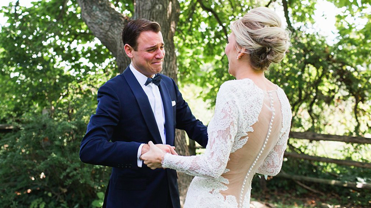 groom in black tux crying as he looks at blonde bride in dress with lace sleeves and cutout back
