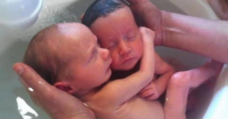 Two newborns cuddling as they're being gently rinsed with water under a faucet.