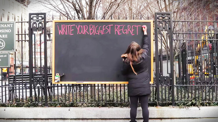 A woman writing "Write your biggest regret" on a large chalkboard outside in NYC.