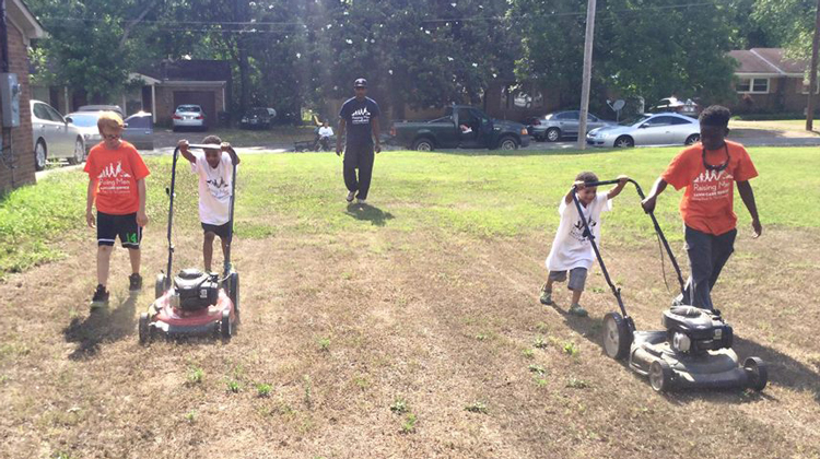 two sets of kids in white shirts mow lawn guided by kids in orange and supervised by man in black