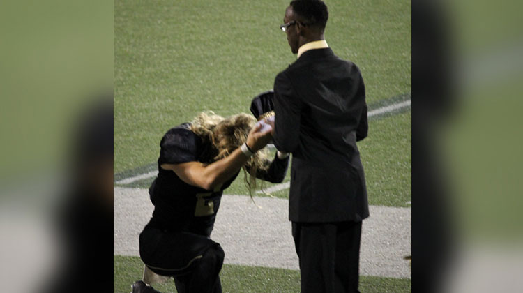 quarterback kneels to present crown to student in black suit