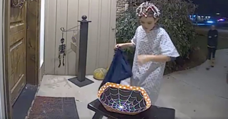 A kid dressed up for Halloween looking into an empty Halloween container on a stranger's front porch.