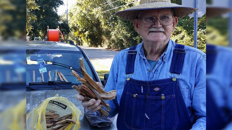 older man holding kindling in straw hat and overalls by car