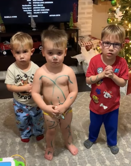 Three toddlers stand in the middle of a room and look guilty, one of them is just wearing his underwear and has marker all over him.