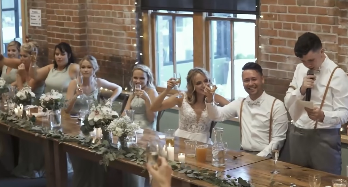 bride and groom with wedding party raising their glasses to toast while one man stands and talks with a microphone