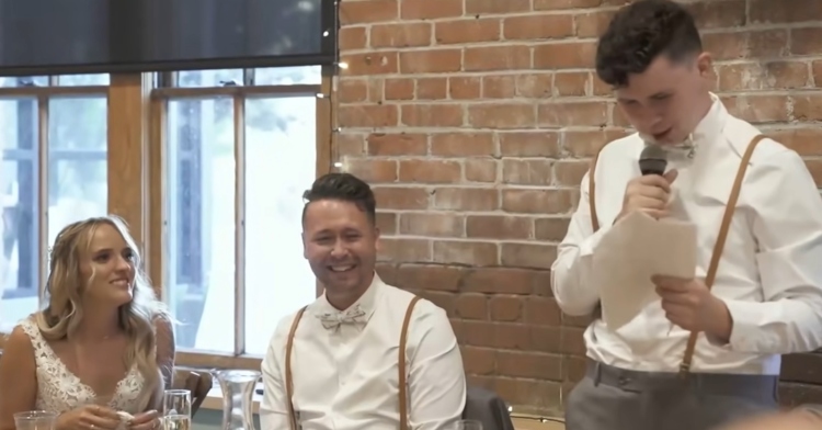 man standing with a microphone and reading from a paper with a groom and bride sitting next to him smiling