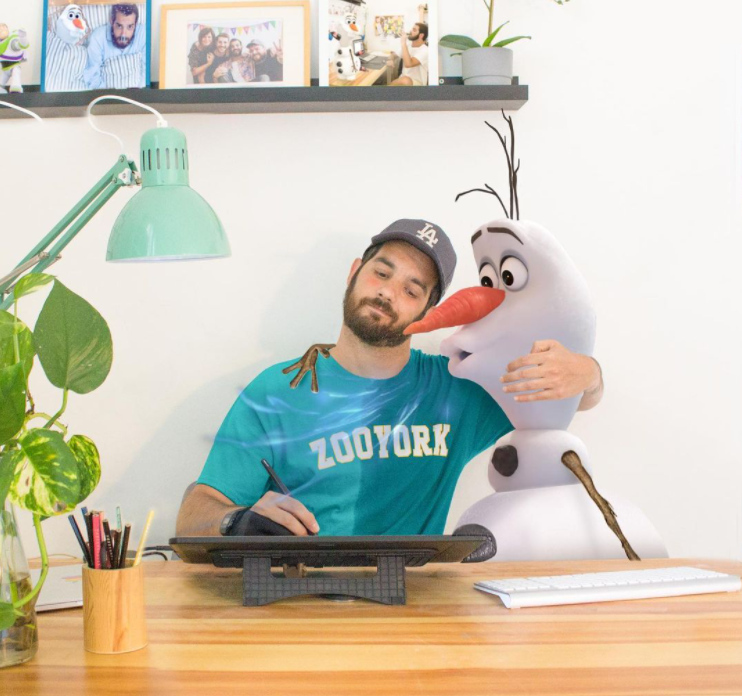 man drawing on a tablet at a desk while he has one arm around olaf from the disney movie frozen