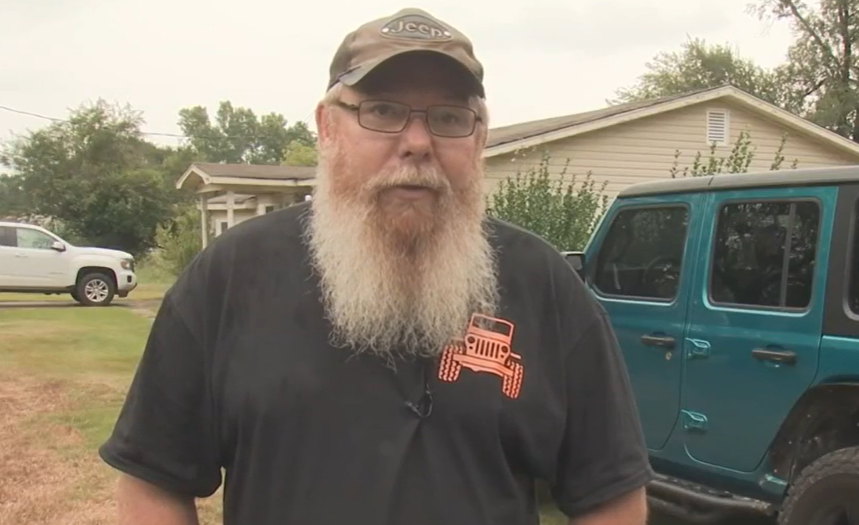 man with beard, glasses, and hat standing in yard next to a jeep