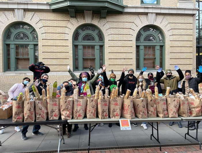 large group of people wearing masks and gloves while standing at tables with bags full of food