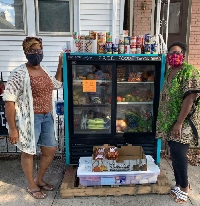 two women wearing masks and posing next to fridge with lots of fresh fruits an vegetables 