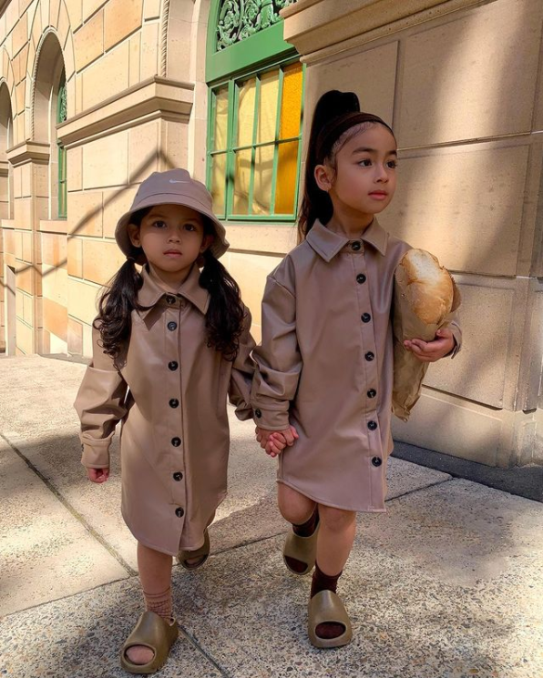 two little girls wearing trench coats while holding hands and walking down the sidewalk