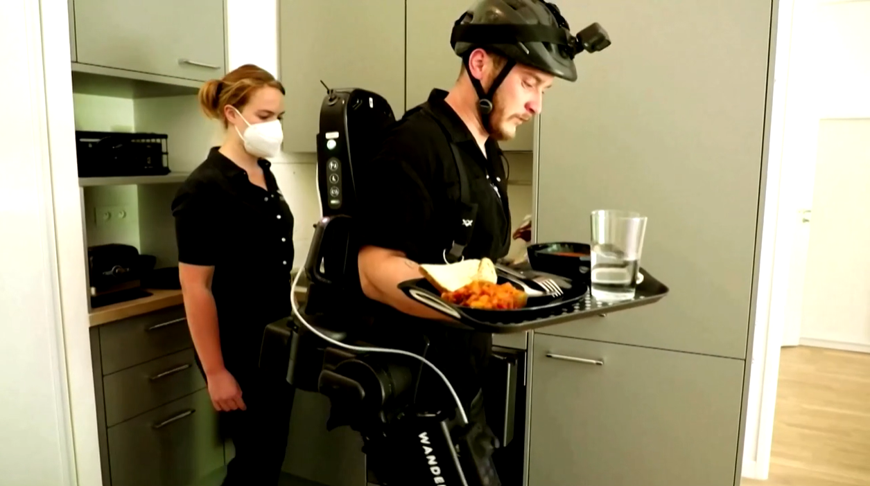 woman in mask walking behind a man walking with robotic exoskeleton while carrying a tray of food