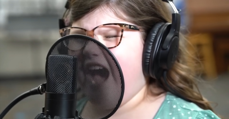 little girl in glasses passionately singing into a microphone while wearing headphones
