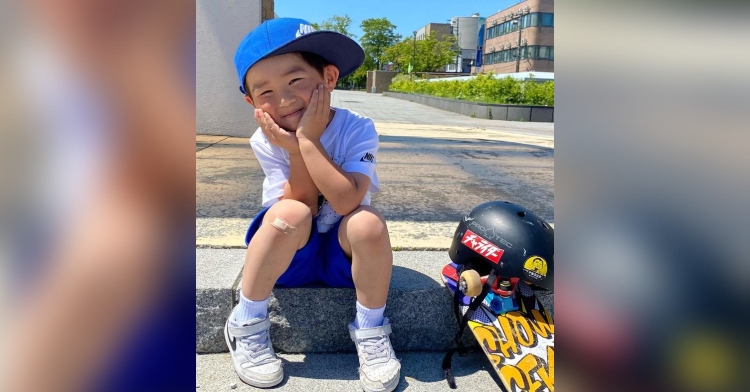 4 year old boy with a band aid on his knee smiling while sitting on a step outside next to a skateboard and a helmet