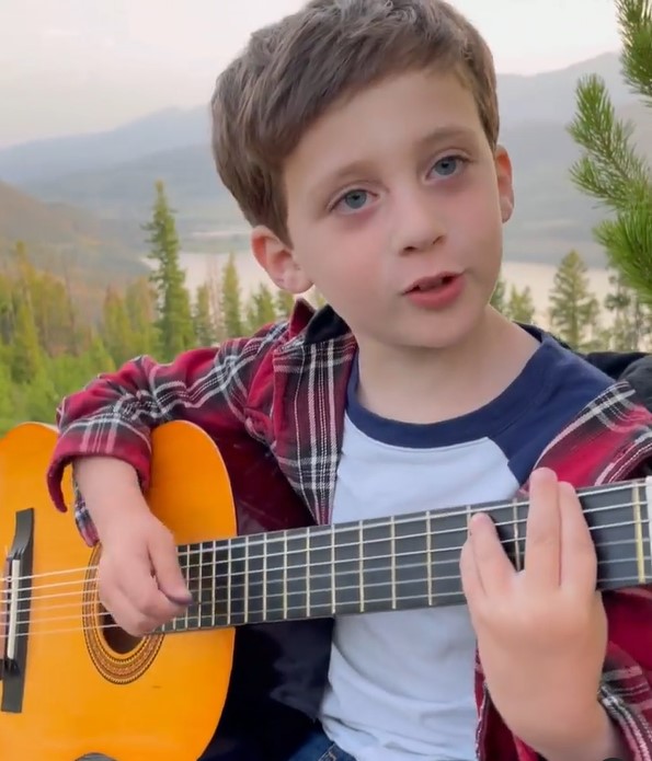5 year old boy sitting outside and singing and playing a guitar with maintains and a lake in the background