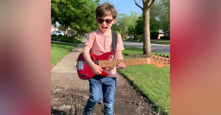 5 year old standing on a sidewalk playing a red guitar while wearing dark sunglasses