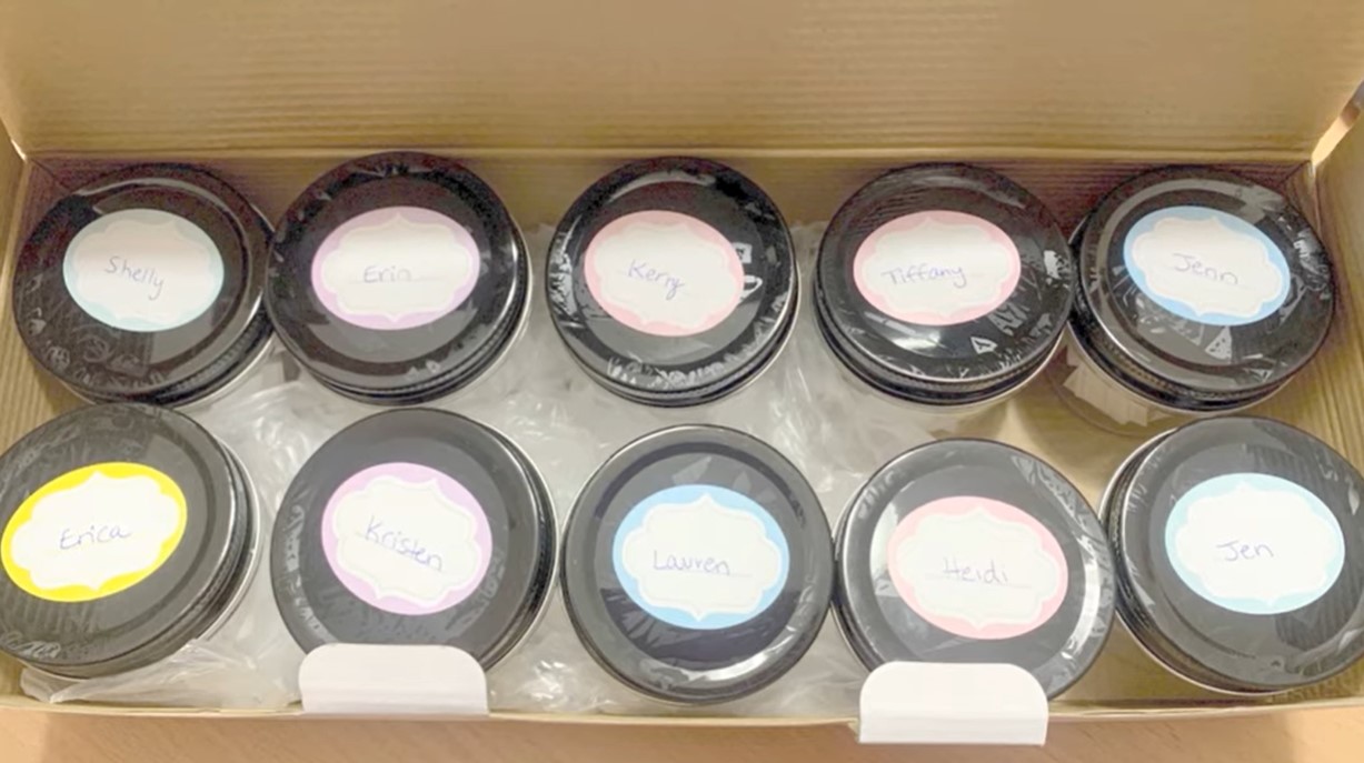 open box full of 10 jars with black lids and stickers with people's names written on them on top of the lids