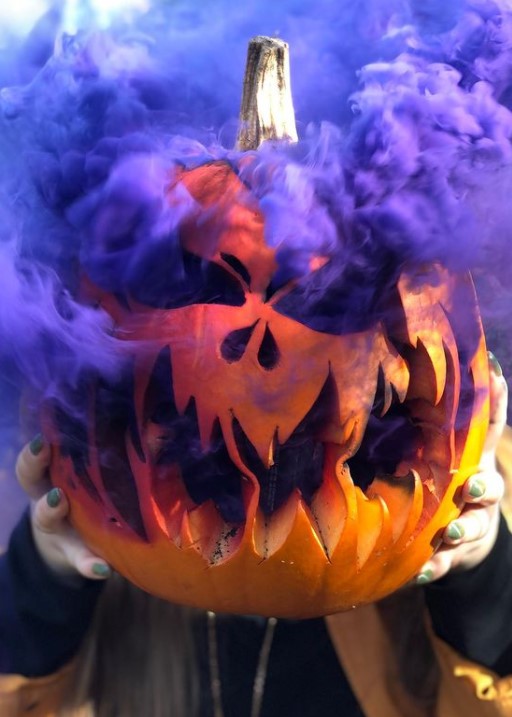 someone's hands holding a carved pumpkin that has purple smoke coming out of its eyes, nose, and mouth