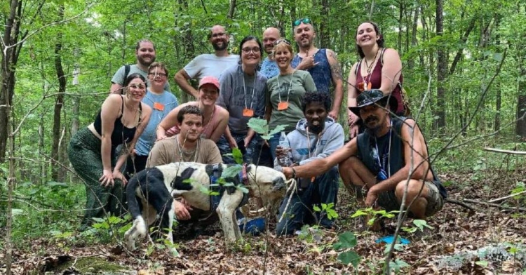 group of thirteen people posing in the woods with a black and white dog