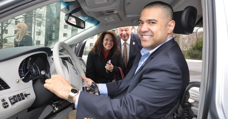 man smiling big in the passenger seat with a man and woman standing smiling and standing outside on the other side of the car with the door open
