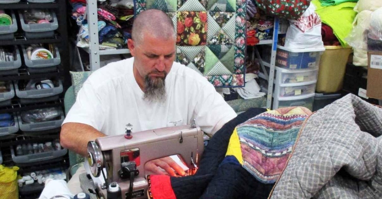 man with shaved head and long beard concentrating while quilting at a table with a quilt and quilting supplies behind him