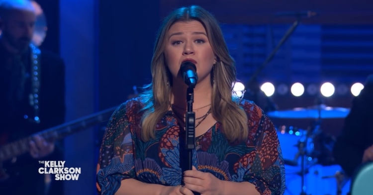 kelly clarkson singing into a microphone on a stand with the kelly clarkson logo at the bottom left corner