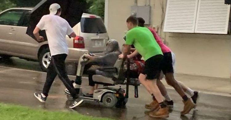 four young men pushing an elderly woman down the road in the rain with an umbrella as they pass a house