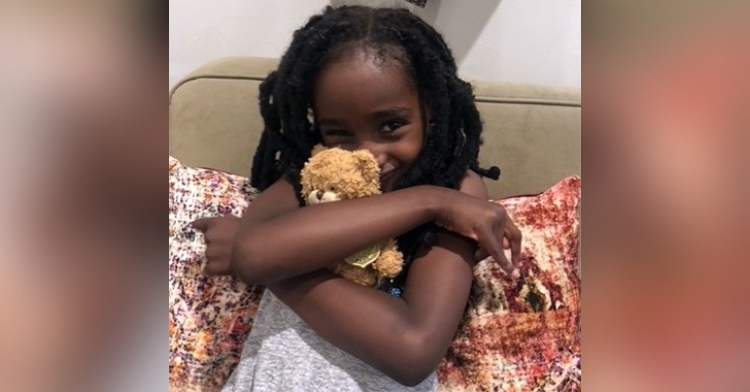 little girl sitting on a couch and smiling big while hugging a small teddy bear