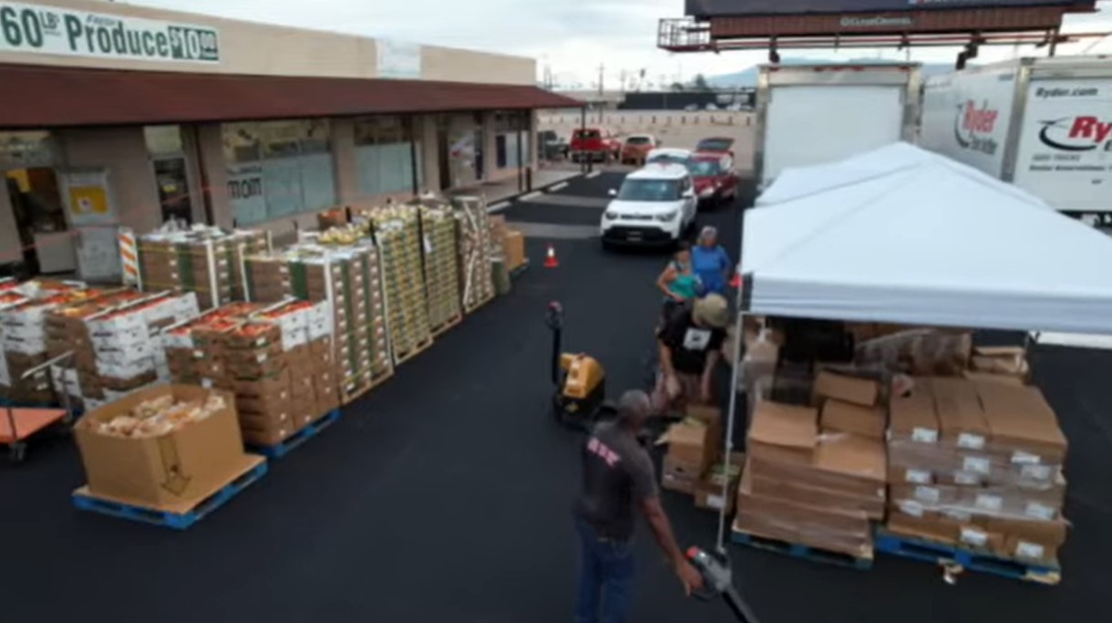 several boxes and crates of produce with people distributing them and a line of cars waiting