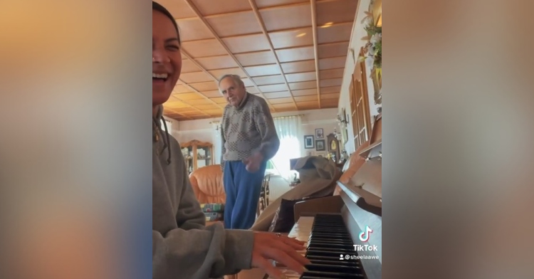 woman smiling while playing the piano with her 93 year old grandpa standing a few feet behind her