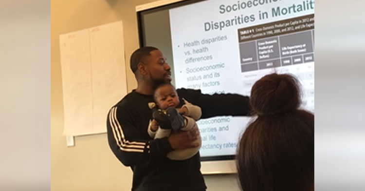 professor holding baby in one arm and pointing at screen with the other