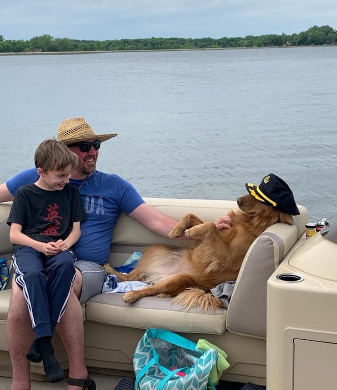 a boat on the water with a man smiling with a kid on his lap while the man pets a golden retriever wearing a captain's hat