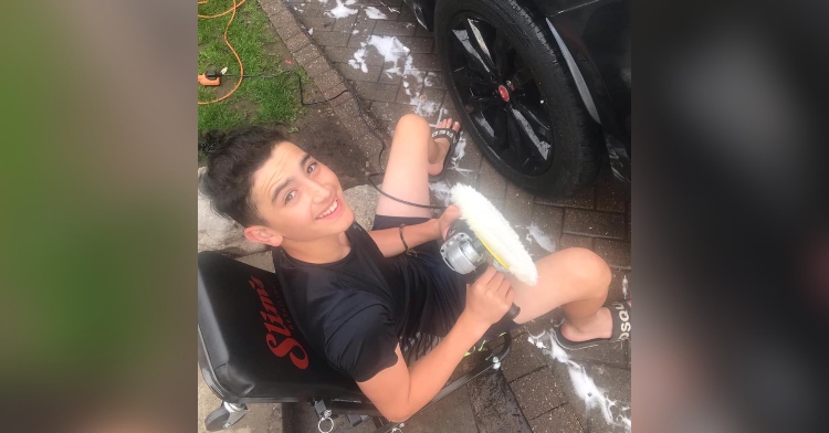 15 year old boy looking up and smiling while sitting on a chair outside as he holds a car buffer and sits near a black vehicle