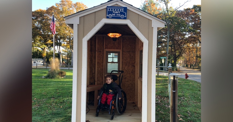 five year old ryder kilam sitting in his wheelchair inside of his bus stop shed that has a sign that reads "ryder's bus stop"