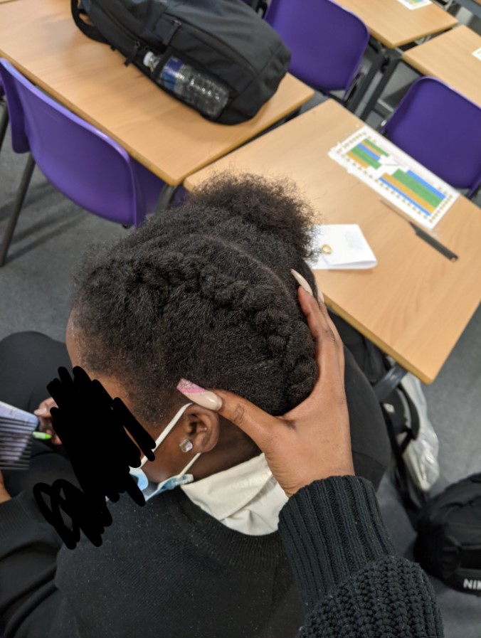 8 year old purity agyeman sitting in a chair as her teacher vanessa sefa uses her hand to show off the little girl's hair which has been styled into two cornrows
