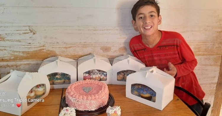 10 year old joaquín nahuel smiling and posing at a table with a pink cake he made along with give boxes containing other baked goods he has made