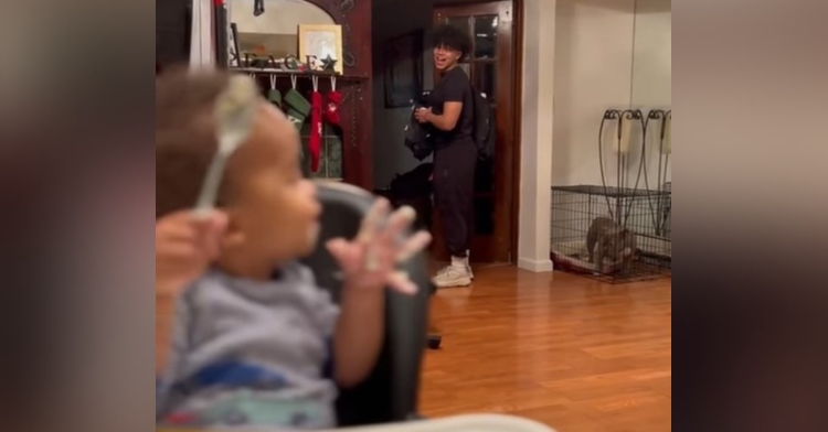 little boy sitting in a high chair eating with a spoon turned around talking to his older brother who is standing at the front door