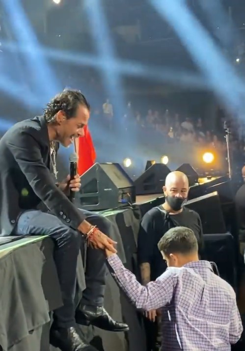 marc anthony sitting on the edge of a stage during a concert as he holds the hand of johnny rivera jr. who is 16 years old and blind