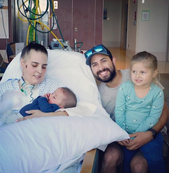 woman named marissa fuentes laying in a hospital bed while holding her baby boy named enzo and her husband, adiran fuentes, and her daughter, ellianna, squat next to the bed