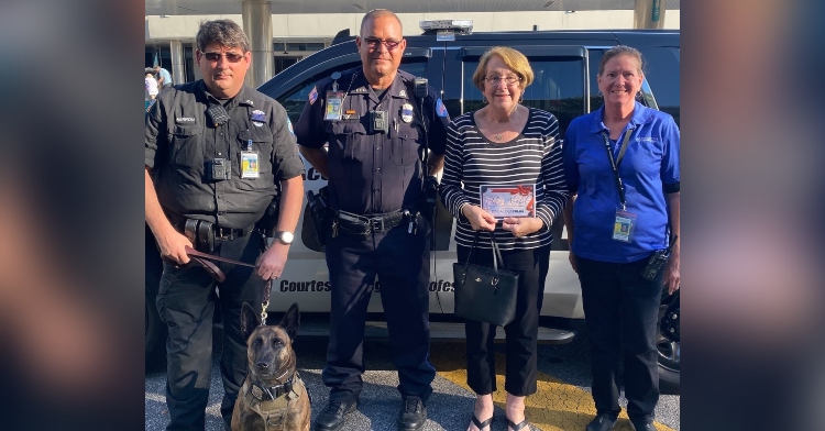 two police officers, a police dog, and a woman from the pensacola international airport smiling and posing with a woman called mrs. ann who is holding the $50 gift card she got from the pensacola police department