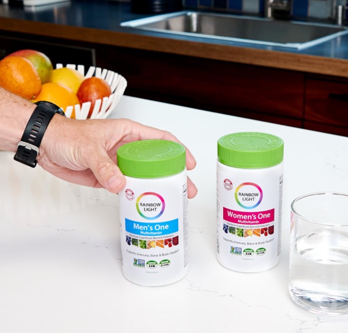closeup of a kitchen counter where someone is grabbing a bottle of rainbow bright men's one multivitamins which are next to a bottle of rainbow bright women's one multivitamins 