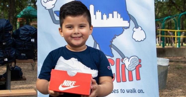 little boy smiling while holding an orange nike box with a white shoe on top as he stands in front of a backdrop for pasos for oak cliff at their shoe drive