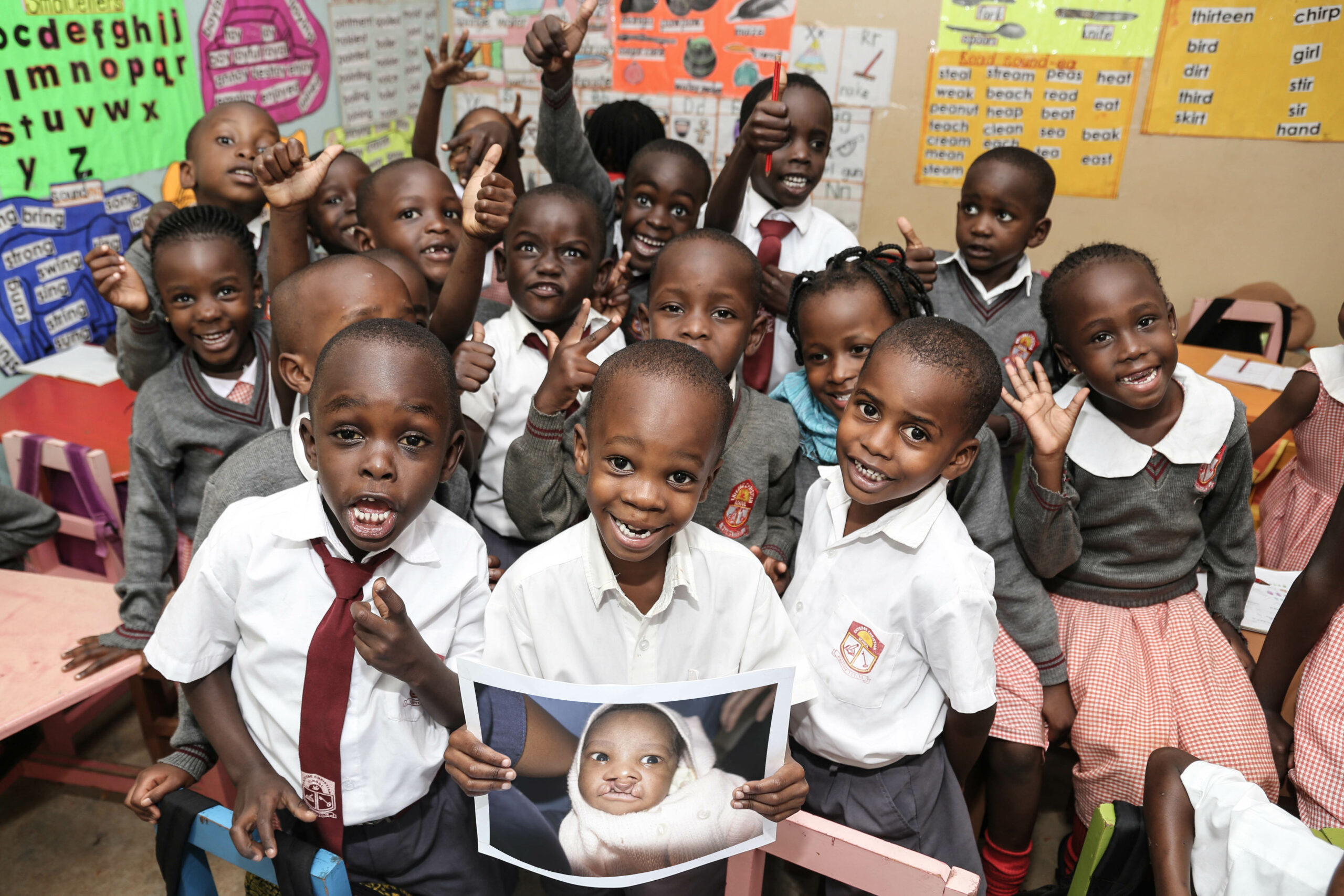 little boy holding picture of himself as a baby with a cleft surrounded by all his classmates in school