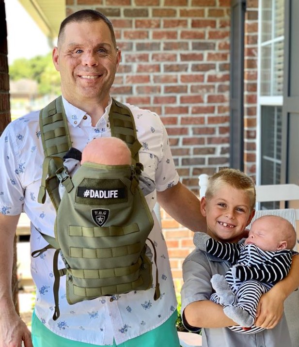man named aaron hale smiling with a baby strapped to his chest while he poses with his son who is holding his baby brother