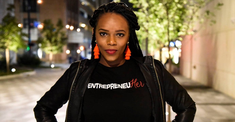 a woman named deidra mayberry posing outside with her hands on her hips as she wear a shirt that reads, "entrepreneuher"