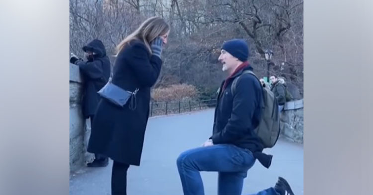 man named brian prentiss down on one knee in central park in nyc to propose as a woman named katie prentiss covers her mouth is surprise