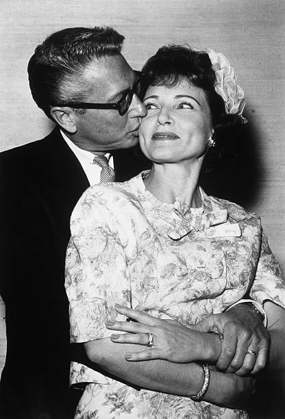 black and white photo of allen ludden with his arms wrapped around betty white from behind as he kisses her cheek and she smiles, looking at him from the corner of her eye