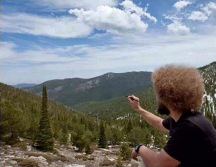 a man who looks like bob ross from the back squatting and holding up a paint brush to look as though he's painting the scenic mountain in the background