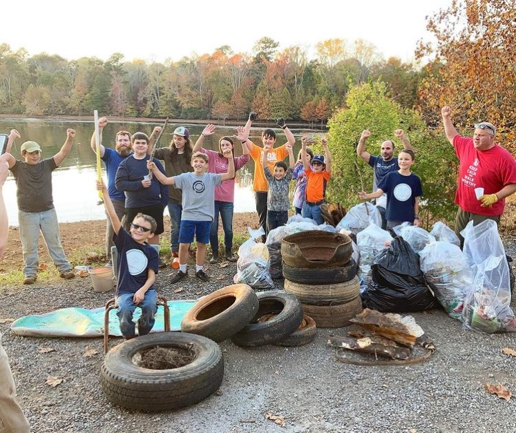 group of kids and adults posing with their hands up around the trash they've cleaned out of the near-by river