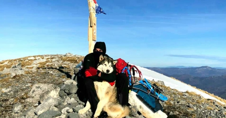 a man named grga brkic and his 8 month old alaskan malamute, north, sitting on a mountain top in croatia in front of a wooden cross